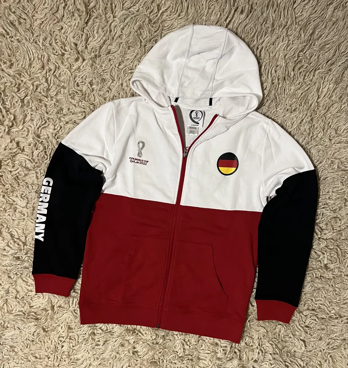 Buzo con capucha y cierre completo, con estampa de FIFA World Cup Qatar 2022 y escudo de Alemania en el frente. Mangas color negro con la palabra "GERMANY" estampada.