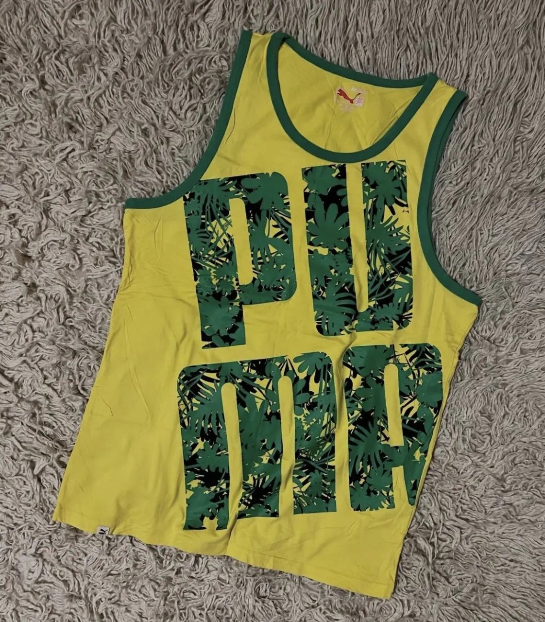 Musculosa amarilla con estampa de hojas en verde y negro en las letras "PUMA". Tiene ribetes verdes en cuello y mangas. Logo de PUMA en la espalda.