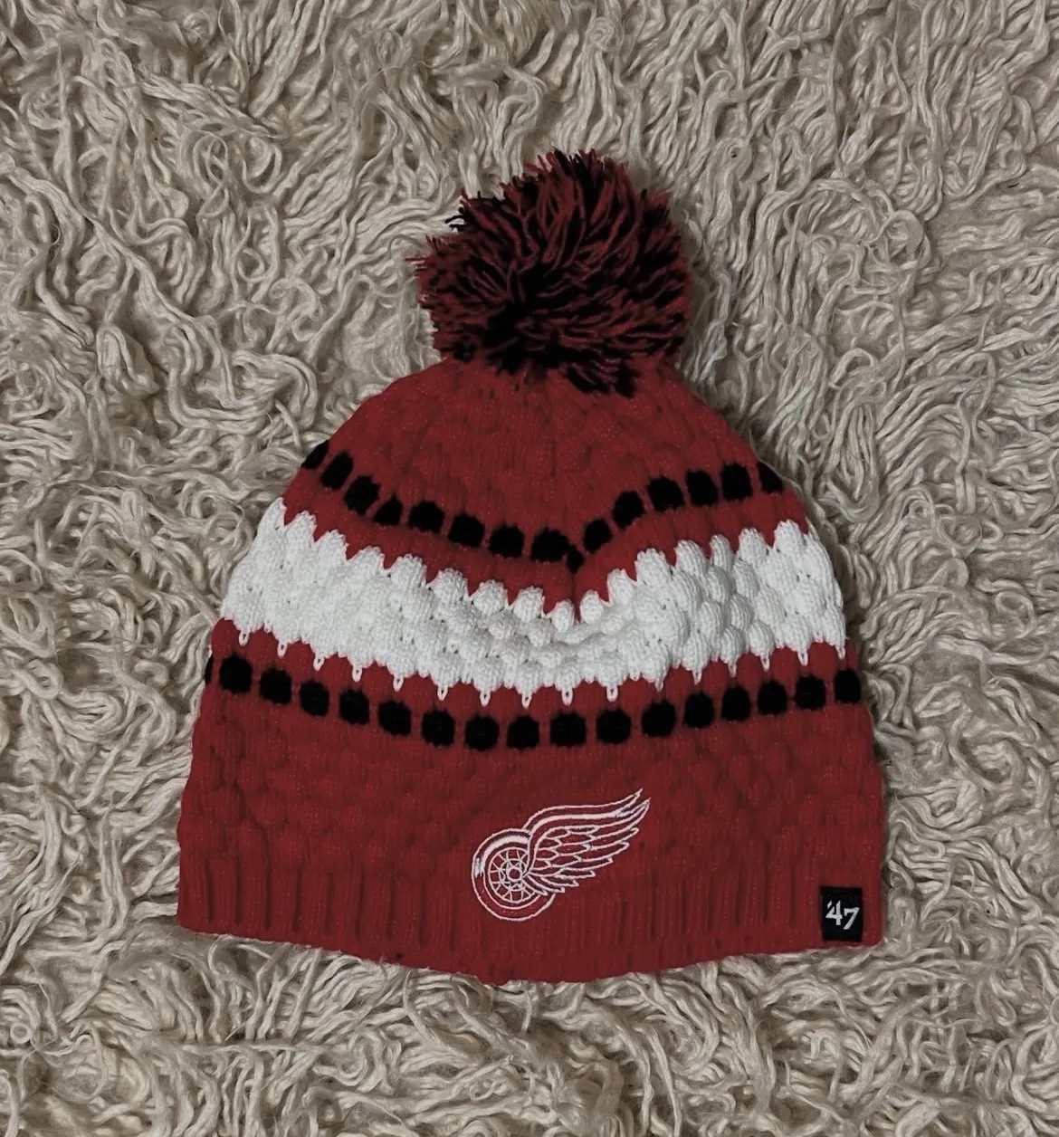 Gorro tejido de lana con pompón, de color rojo, blanco y negro. Presenta un diseño de franjas y el logo de un equipo deportivo bordado.