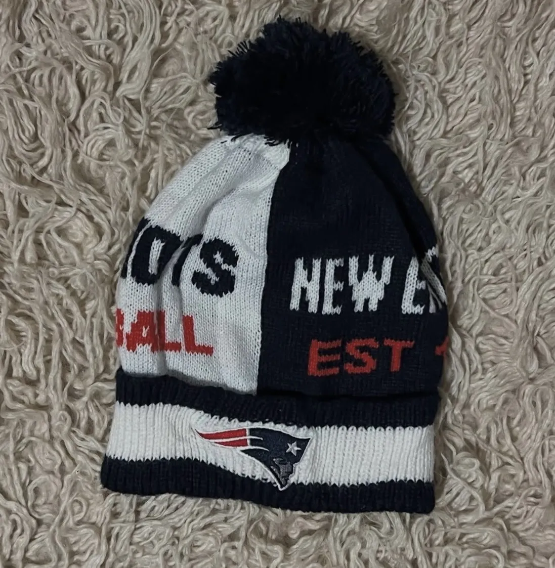 Gorro tejido con pompon, de los New England Patriots. Tiene el logo del equipo bordado y el nombre del equipo y "FOOTBALL" escrito.