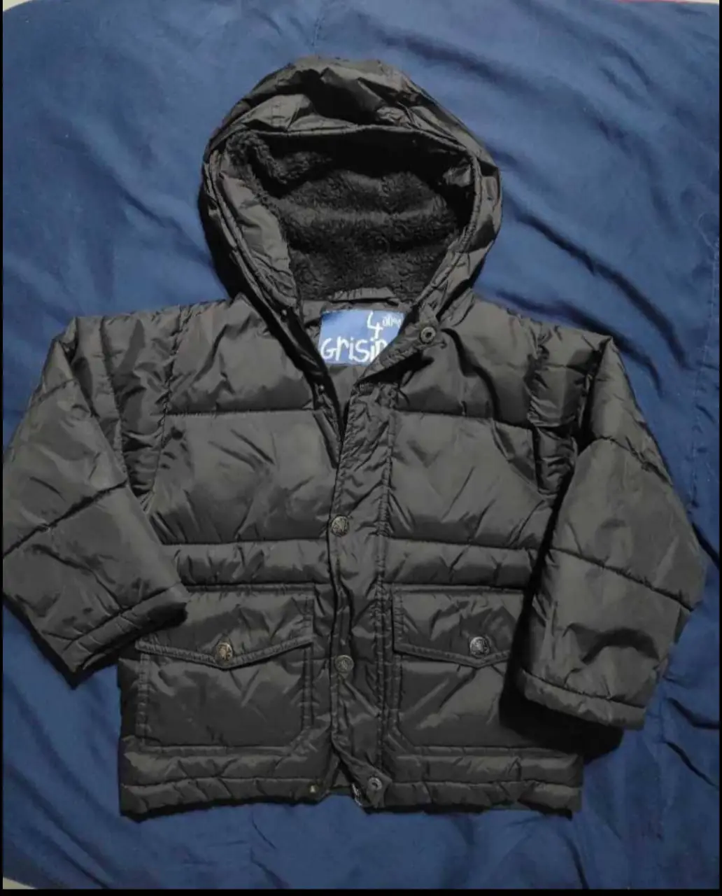 Campera negra acolchada para niño, con capucha forrada en piel sintética. Tiene dos bolsillos delanteros y cierre con botones. Ideal para el invierno.