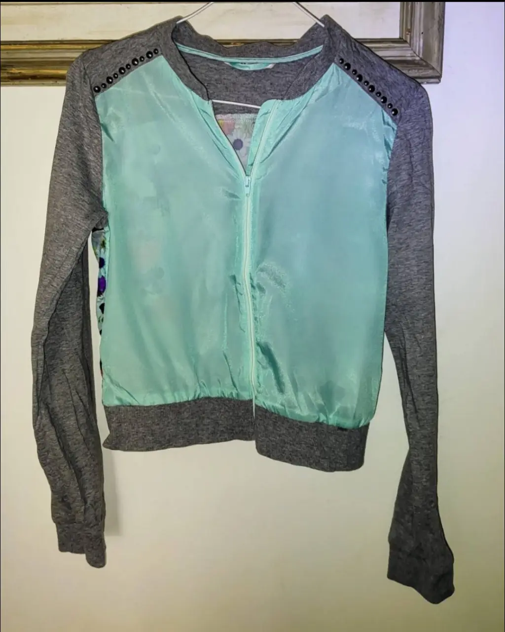 Buzo con cierre para mujer, con mangas grises y frente celeste. Detalles de tachas en los hombros y estampado de caballo con flores en la espalda. Ideal para un look casual y cómodo.