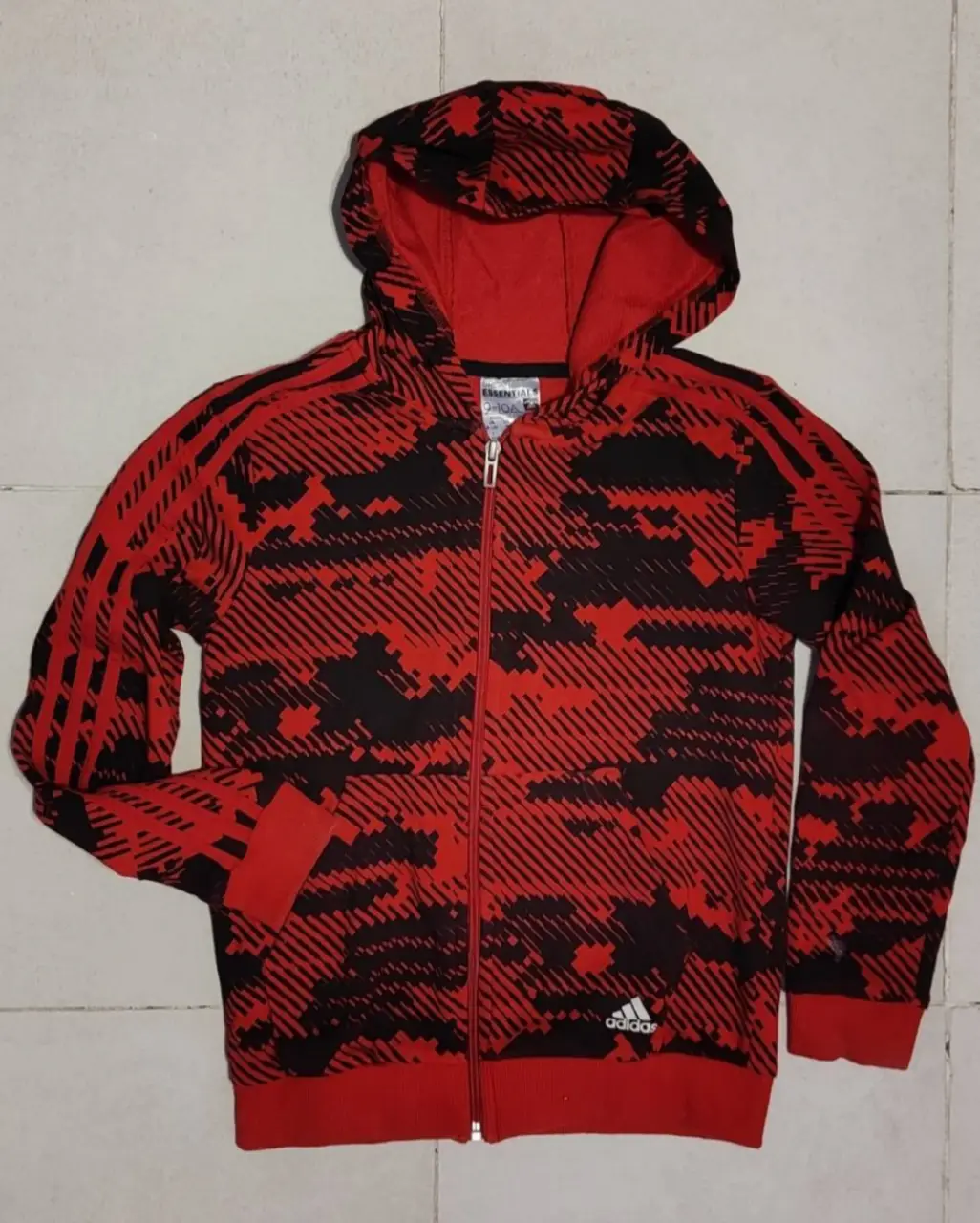 Buzo con cierre Adidas Essentials con estampado camuflado en rojo y negro. Ideal para un look deportivo y casual.