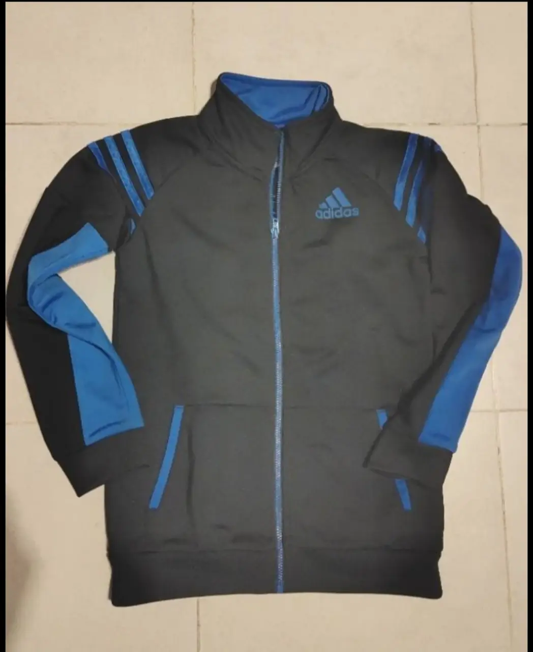 Buzo deportivo Adidas con cierre, color gris con detalles en azul. Ideal para actividades deportivas o uso casual.
