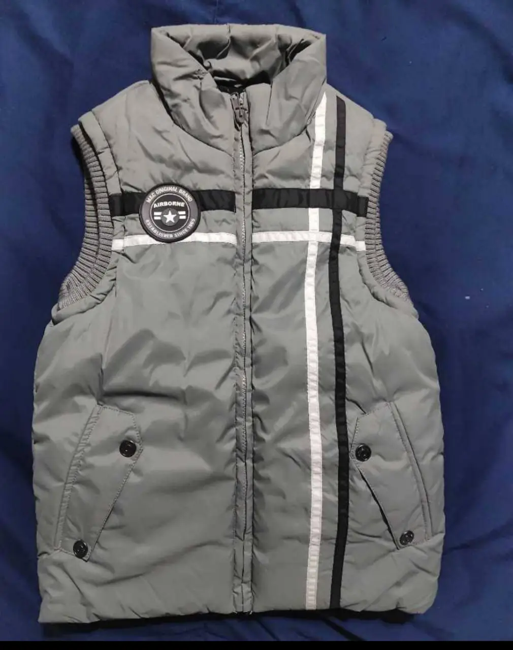 Chaleco gris acolchado para niño, con detalles en negro y blanco. Tiene cierre y dos bolsillos delanteros. Incluye un parche con el logo de la marca.
