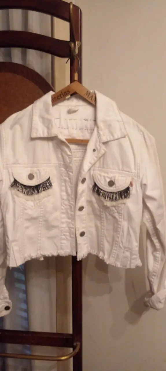Campera de jean blanca customizada con detalles de pestañas en los bolsillos y apliques en la espalda. Corte cropped y detalles desflecados.