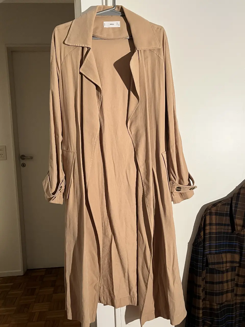 Trench coat largo color crudo de la marca MNG. Ideal para un look elegante y versátil. Confeccionado en un tejido ligero y fluido, este trench presenta un diseño clásico con solapas amplias, cinturón para ajustar la cintura y mangas largas con detalles en los puños. Perfecto para usar en cualquier ocasión.