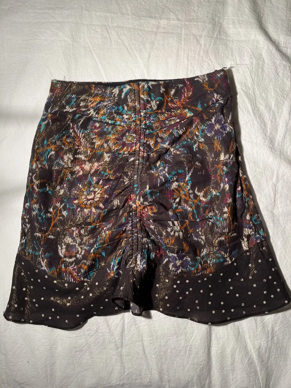 Pollera corta con estampado abstracto en tonos marrones, naranjas, azules y blancos. Detalles de fruncido en la parte delantera y borde inferior con lunares blancos sobre fondo negro. Ideal para un look bohemio y con estilo.