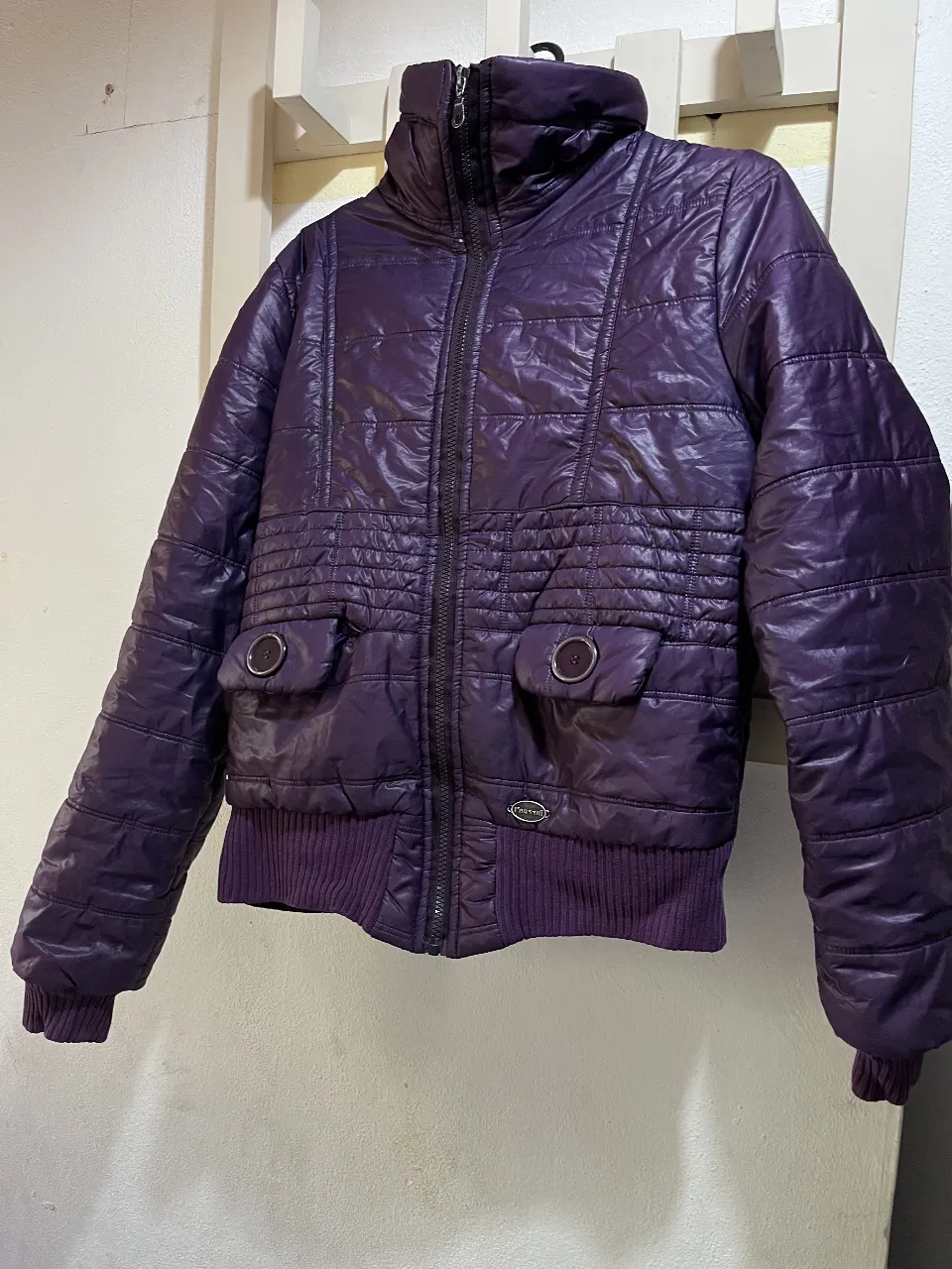 Campera acolchada morada con cuello alto y cierre completo. Posee dos bolsillos delanteros con tapa y botón. Puños y cintura de ribb. mínimo detalle que se ve en la última foto.