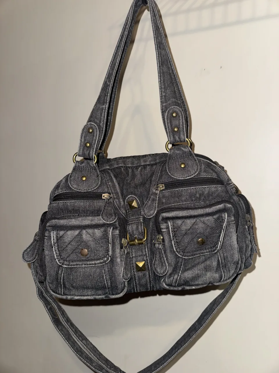 Bolso de jean gris oscuro con múltiples bolsillos frontales y detalles de tachas doradas. Posee dos asas cortas y una correa larga desmontable. El interior está forrado y cuenta con dos compartimentos con cierre.