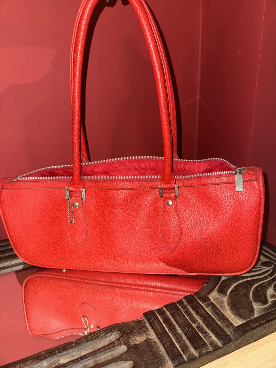 Bolso de cuero rojo con cierre y dos asas largas. Tiene un detalle de cadena colgando de una de las trabas del cierre.