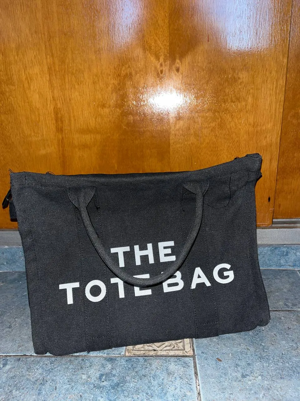 Bolso negro de tela con la inscripción "THE TOTE BAG" en blanco. Tiene dos asas cortas y una correa larga desmontable.
