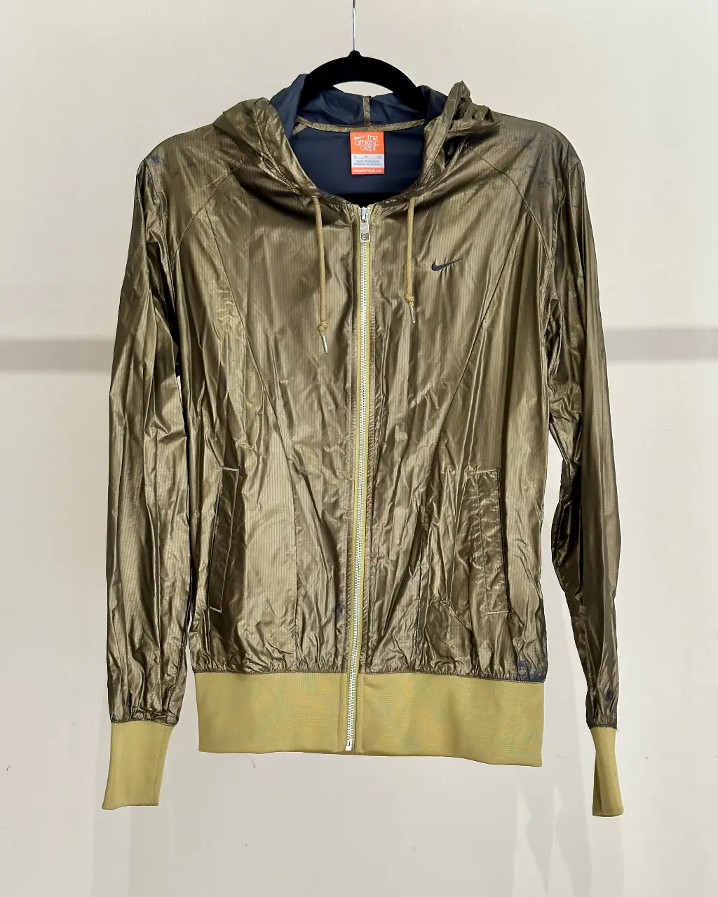 Campera deportiva impermeable Nike dorada con capucha y cierre. Ideal para running o actividades al aire libre. Detalles de rib al tono y logo de la marca. Tiene detalles de desgaste en la zona del hombro por estar colgada.