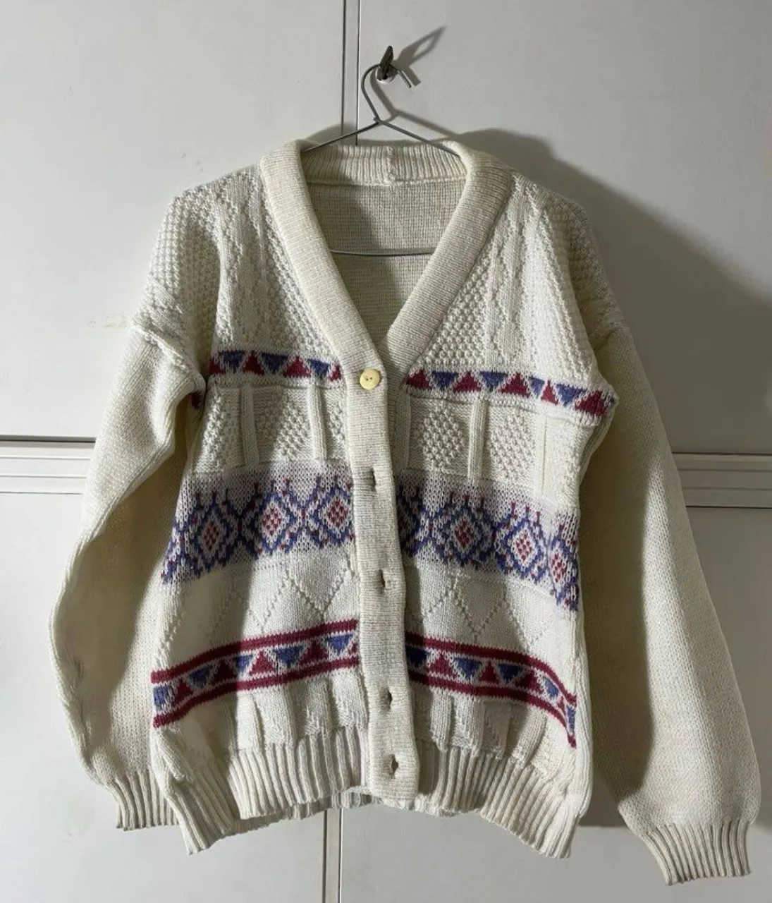 Cardigan de tejido de punto color crudo con detalles de rombos y líneas en color azul y rojo. Posee botones al frente y cuello en V.