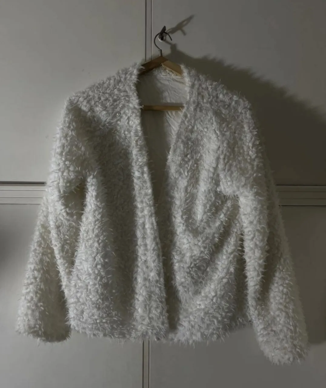 Cardigan blanco de pelo sintético con detalles de perlas pequeñas en el frente. Diseño abierto sin botones ni cierre.