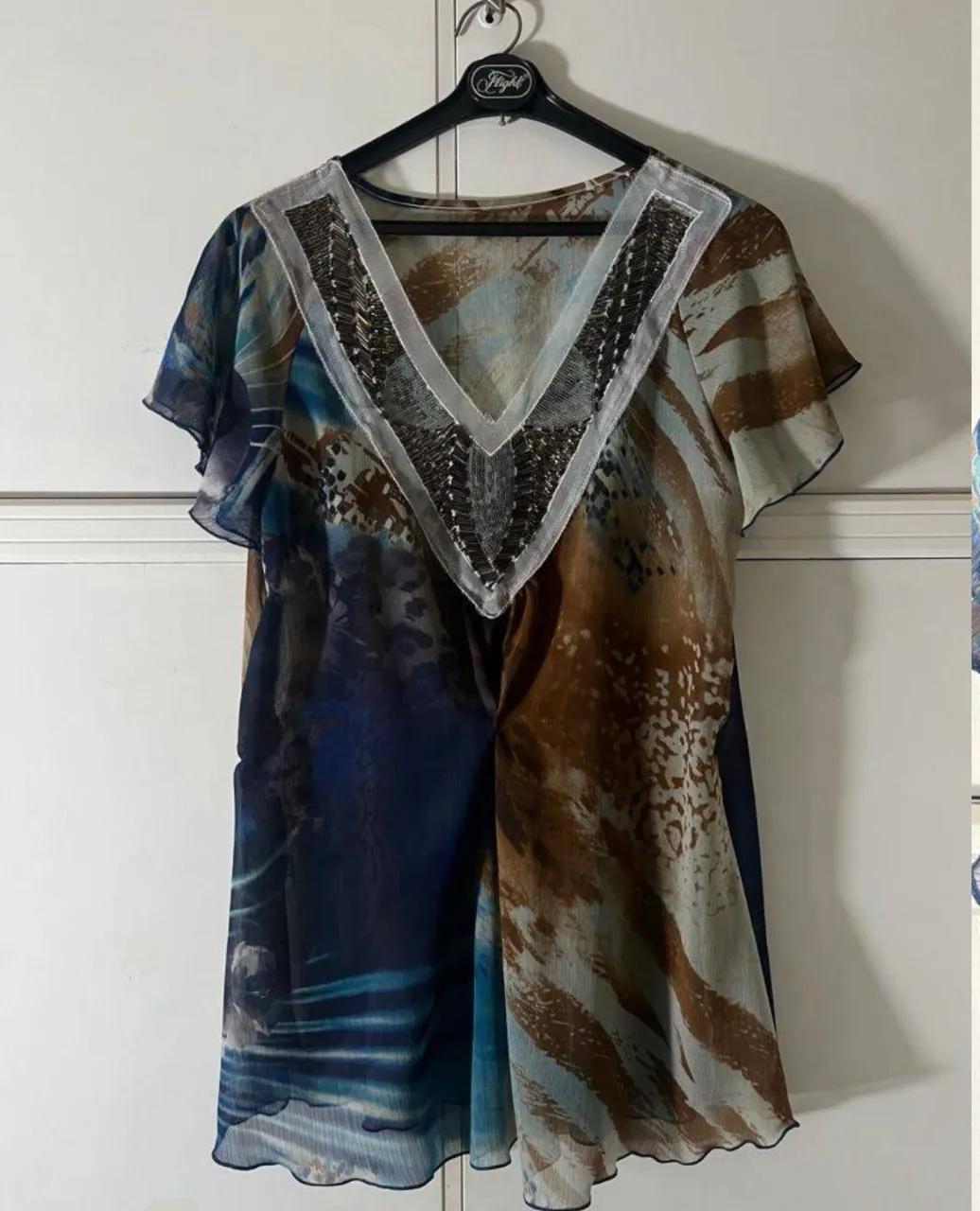 Remera estampada con cuello en V y detalles de pedrería y mostacillas. Posee mangas cortas con volados y terminación ondulada en el borde inferior.