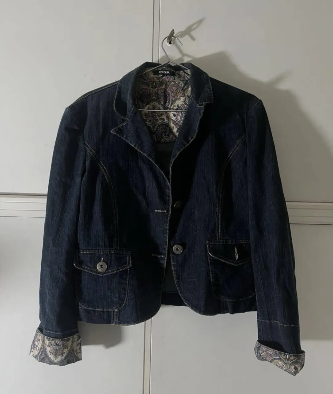 Blazer de jean azul oscuro con forro estampado floral y de paisley en tonos rosas, lilas y amarillos. Posee dos bolsillos frontales con solapa y botones.