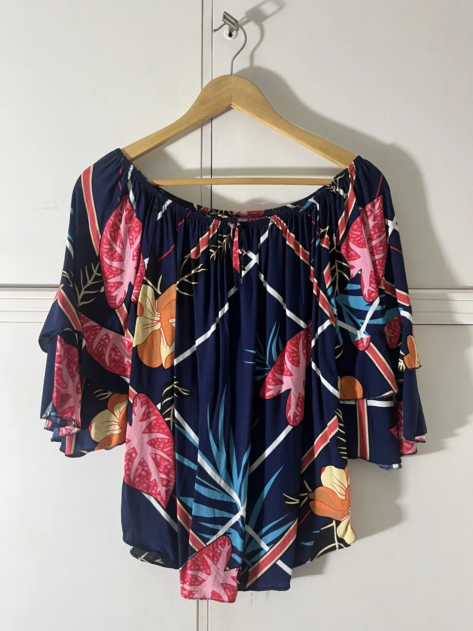 Blusa azul con estampado de hojas y flores en tonos rosa, naranja y celeste. Posee cuello con elástico y lazo, mangas acampanadas de tres cuartos y volados.