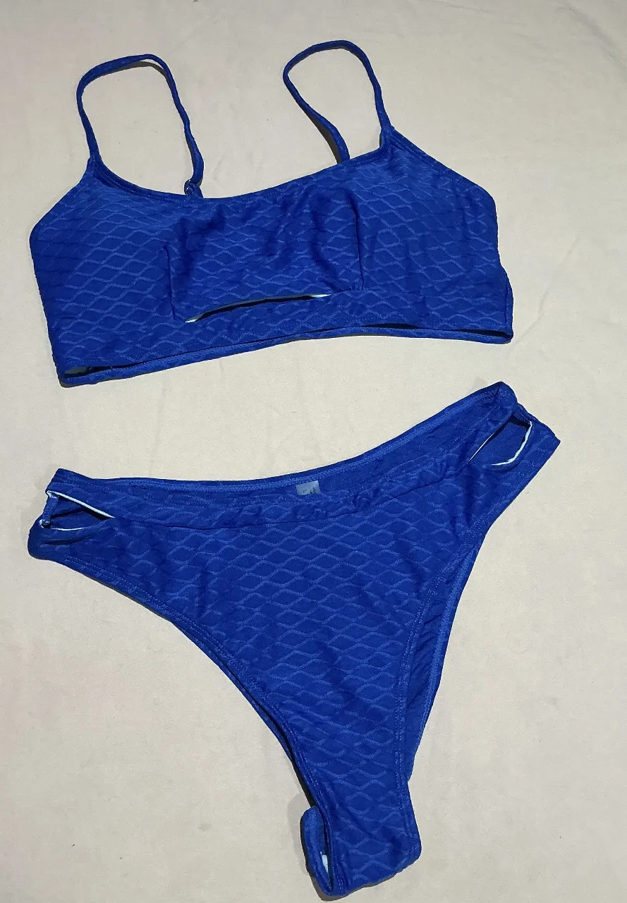 Conjunto de bikini azul vibrante con un diseño texturizado de rombos. El corpiño es tipo top deportivo con breteles finos y un detalle de corte en la parte inferior. La bombacha es tiro alto con recortes laterales y detalles en blanco en los bordes. Ideal para disfrutar del sol y el mar con estilo.