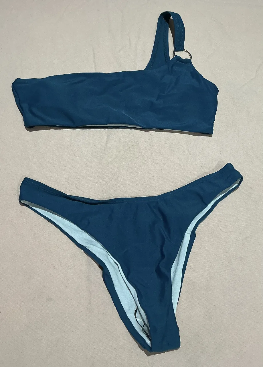 Conjunto de bikini de dos piezas, color azul petróleo. El corpiño es de un solo hombro con un detalle de aro metálico en la tira. La bombacha es estilo colaless.