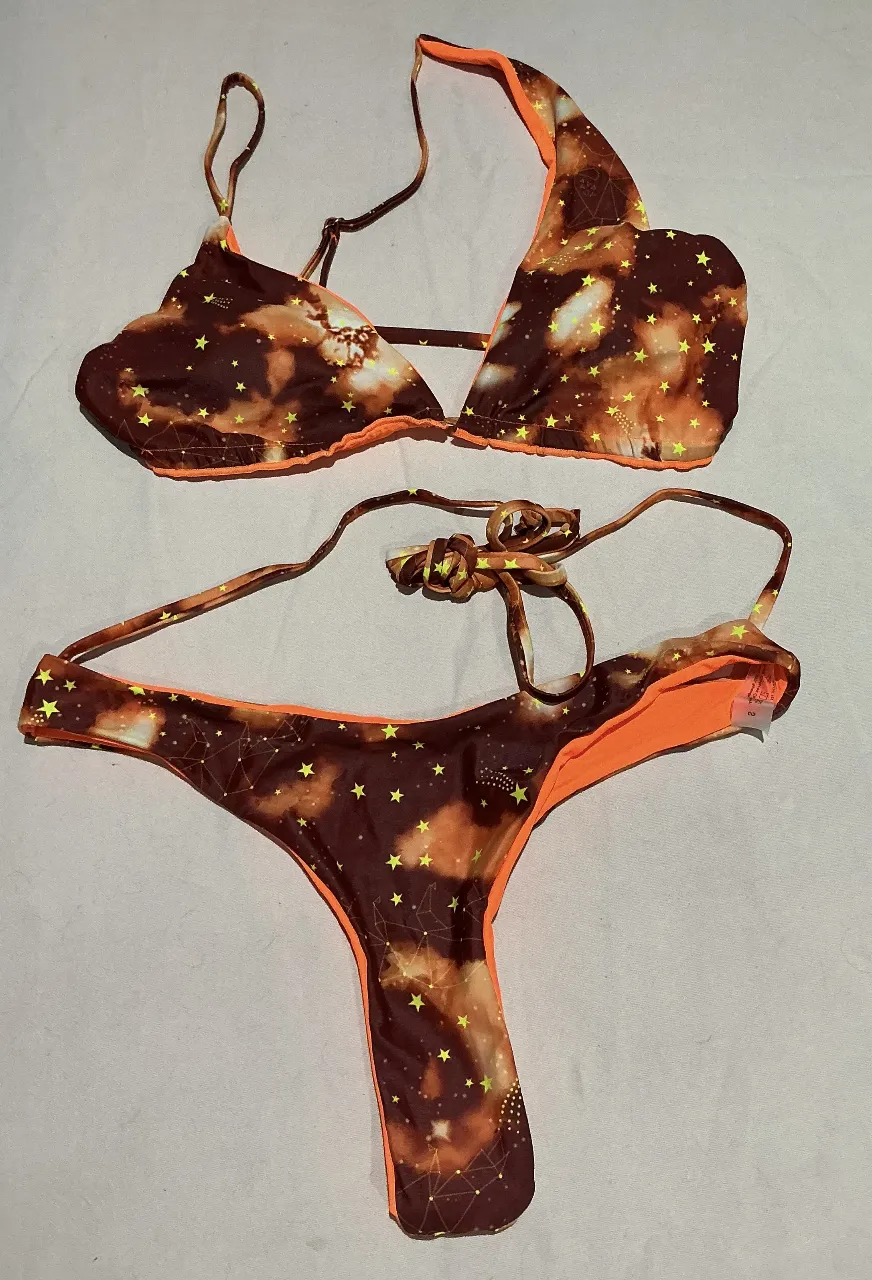 Bikini de dos piezas con estampado galáctico y estrellas amarillas. El corpiño es triangular con tiras finas y el culotte es estilo colaless. Los bordes son de color naranja vibrante.