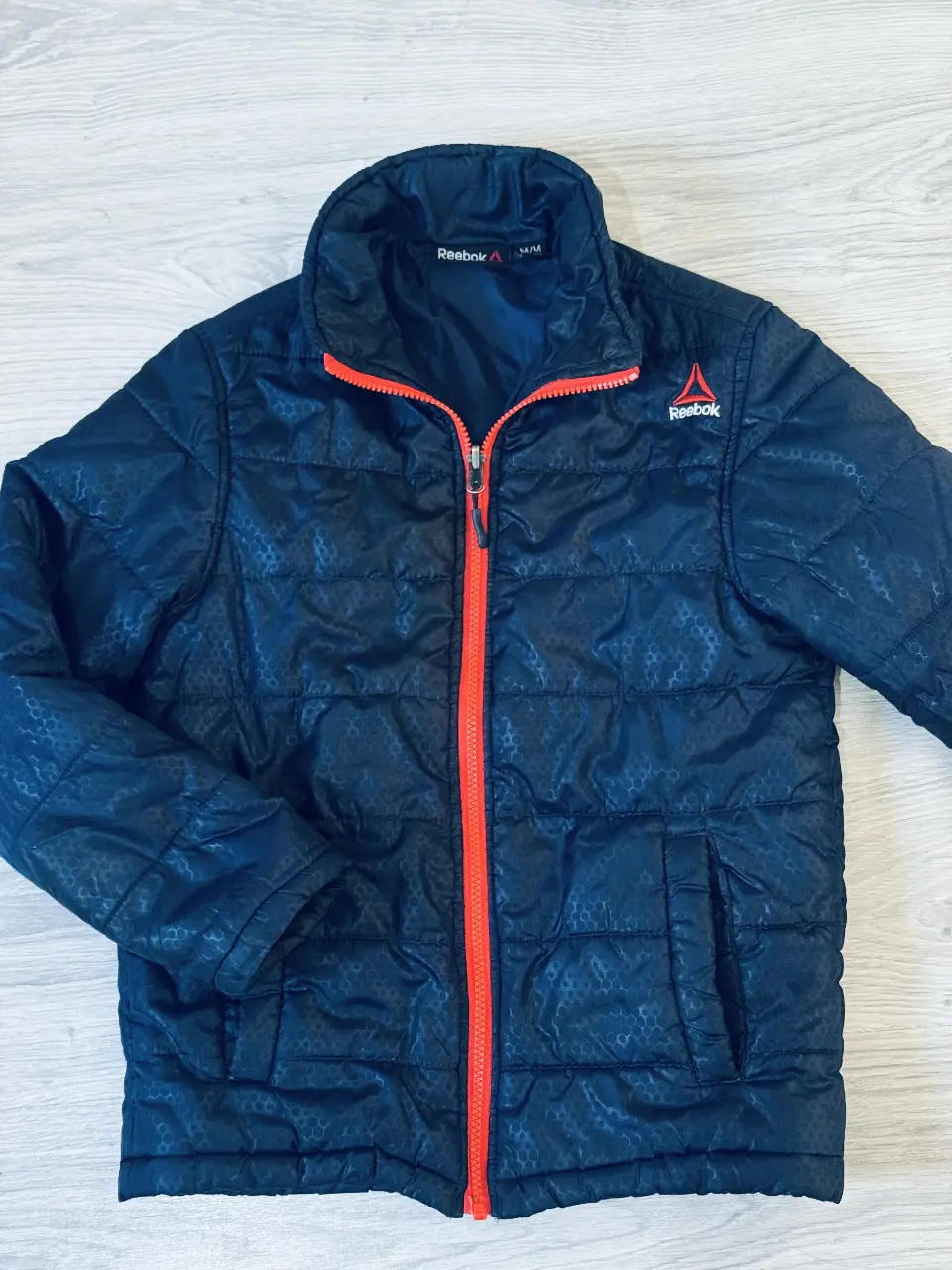 Campera deportiva para niño Reebok, azul marino con detalles en naranja. Con cierre frontal y logo de la marca. Ideal para actividades al aire libre.
