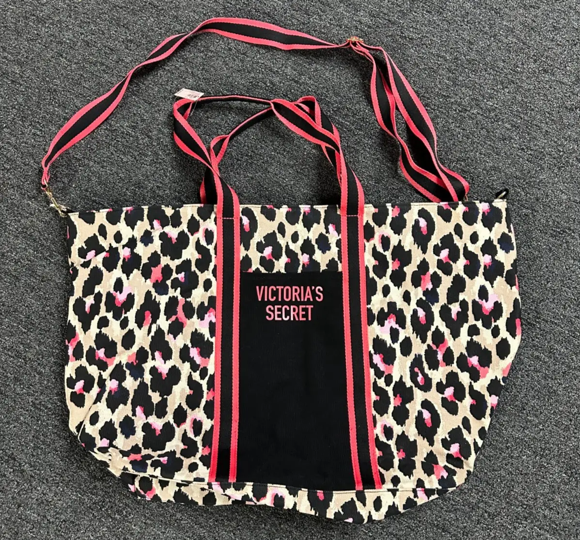 Bolso tote de Victoria's Secret con estampado de leopardo. Cuenta con manijas y correa para el hombro a rayas rosas y negras, y un bolsillo frontal negro con el logo de la marca.