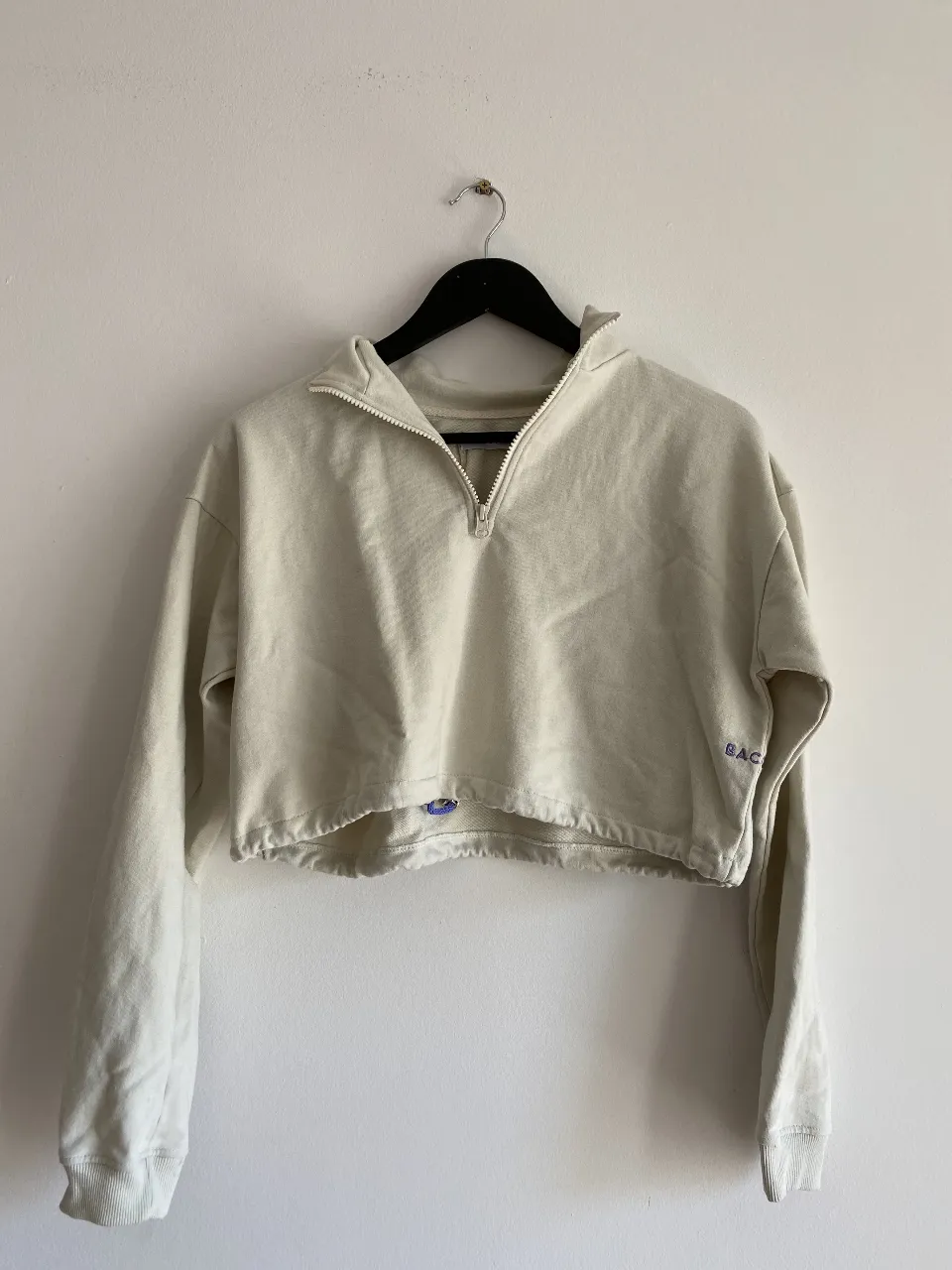 Buzo crop de color crudo con cierre y detalle de bordado. Ideal para un look casual y moderno.