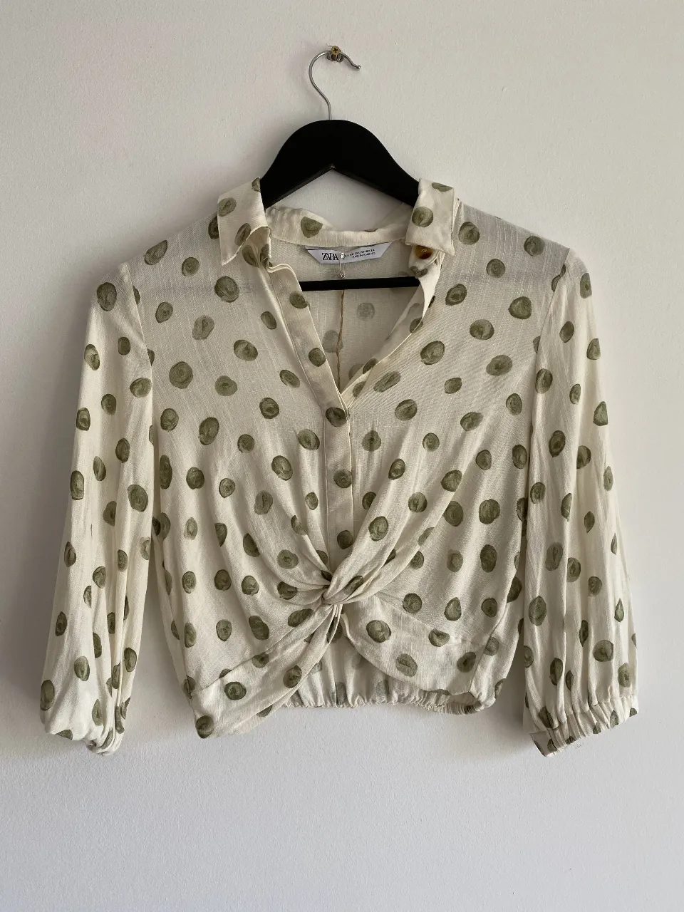 Blusa de mujer con estampado de lunares verdes sobre fondo blanco. Diseño con cuello camisero, botones y detalle de nudo en la cintura. Ideal para un look casual y moderno.