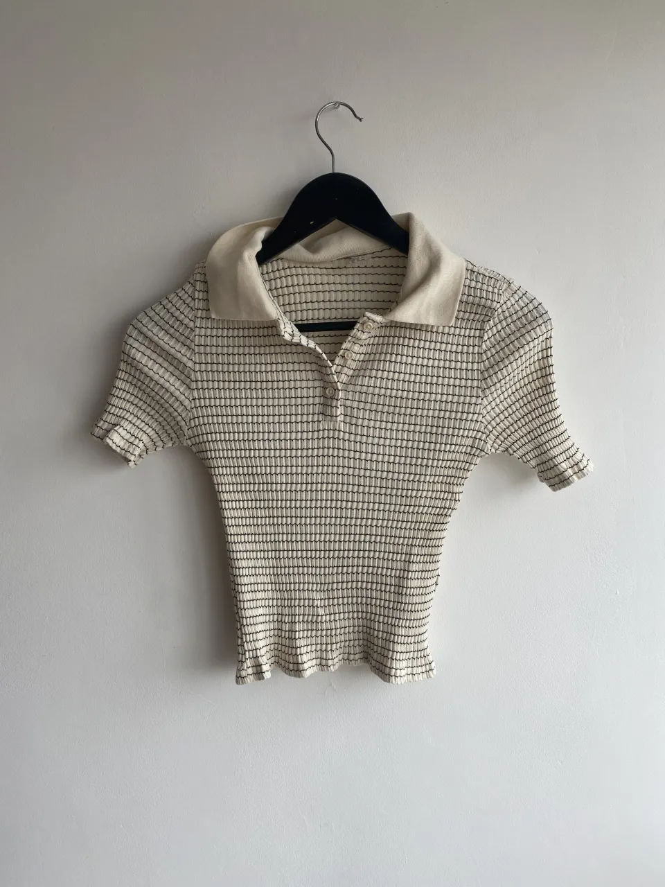 Remera polo de manga corta con textura acanalada y estampado de finas rayas verticales en tonos beige, blanco y marrón. Cuello camisero y botones al frente. Ideal para un look casual y chic.