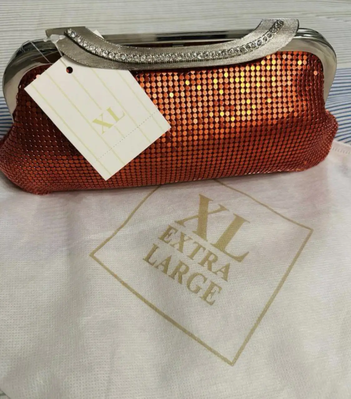 Cartera de mano XL, color rojo metalizado, con detalles de strass en el borde superior, cadena y herrajes plateados. Ancho superior 18 cm y ancho inferior 25 cm. interior de raso rojo con bolsillo tarjetero. Incluye bolsa protectora.