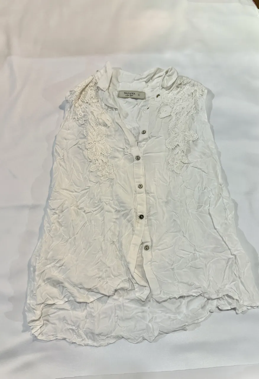 Blusa blanca de mangas cortas con detalles de encaje en los hombros y escote en V. Posee botones al frente y un diseño arrugado que le da un toque bohemio y chic. Perfecta para un look casual y fresco.