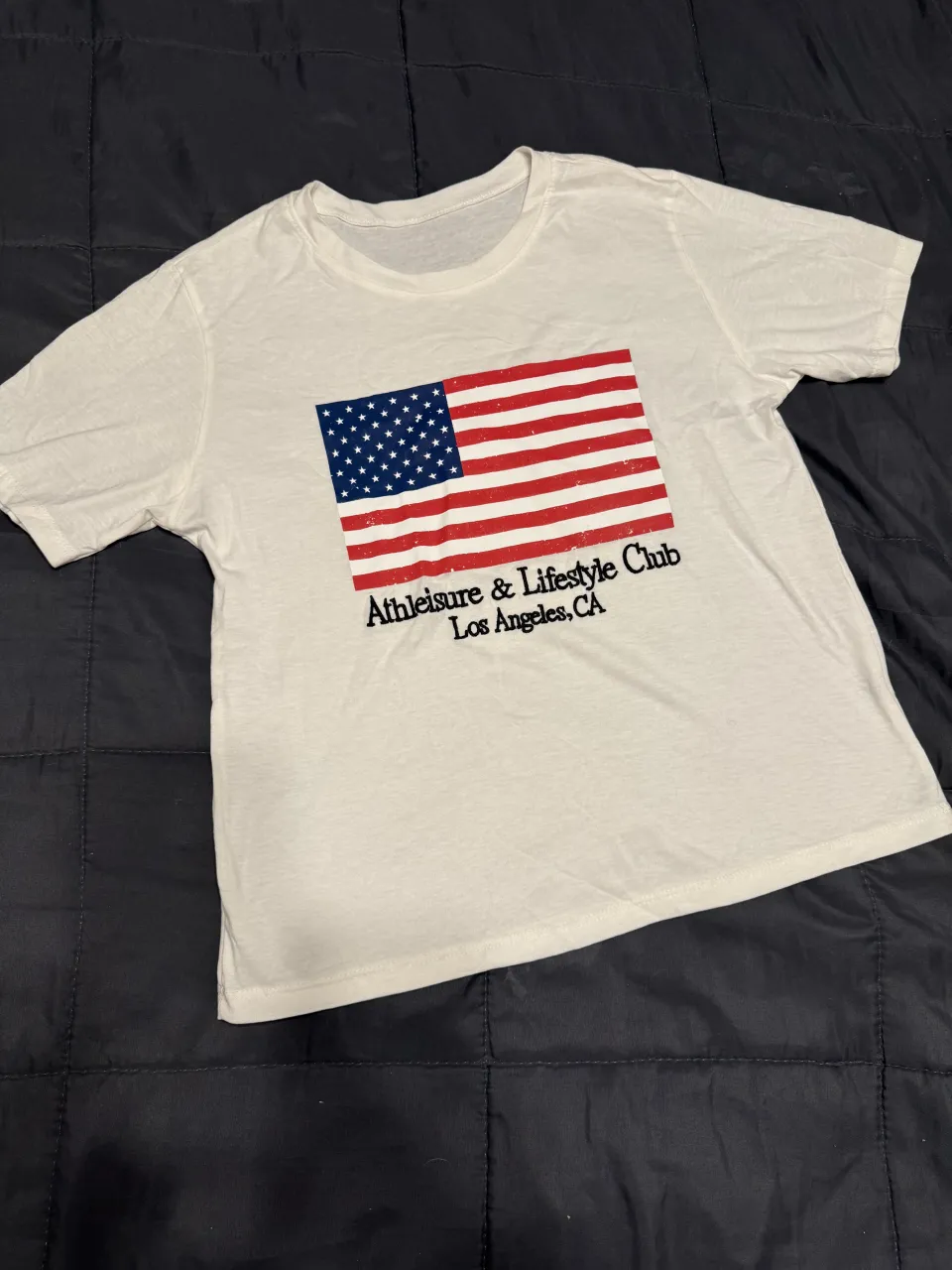 Remera blanca con estampa de la bandera de Estados Unidos y texto "Athleisure & Lifestyle Club Los Angeles, CA". Ideal para un look casual y relajado.