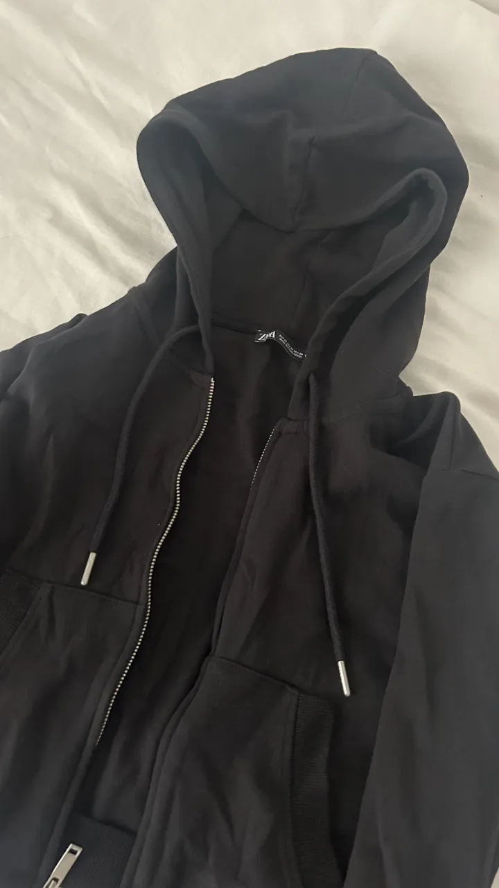 Buzo ZARA negro crop (corto) con capucha y cierre. Tiene bolsillos frontales y cordones en la capucha.