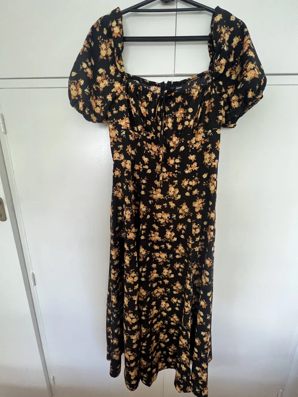 Vestido negro con estampa floral en tonos amarillos y naranjas. Escote cuadrado con detalle de lazo en el frente. Mangas cortas abullonadas. Largo hasta la rodilla con tajos laterales y abertura a los lados