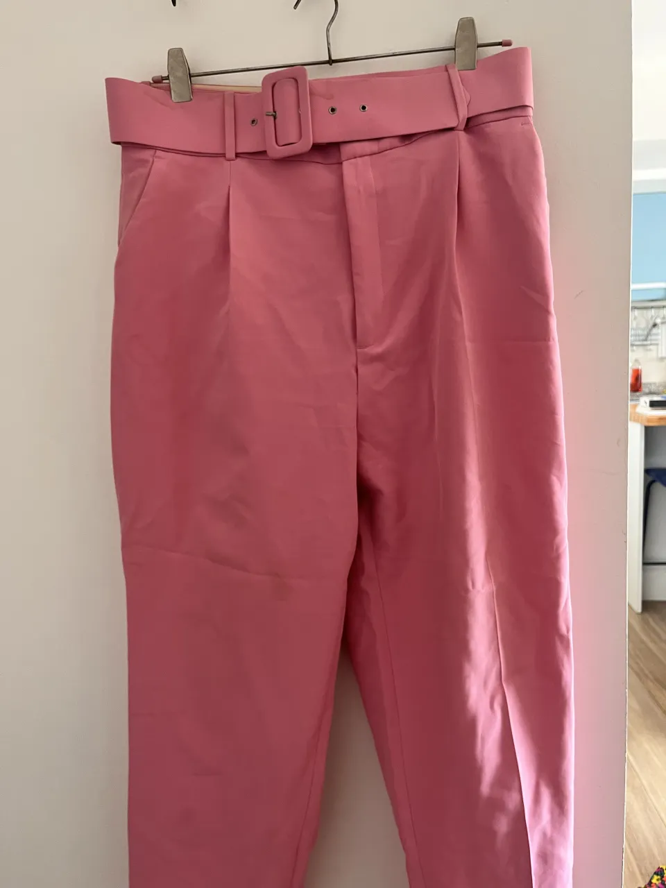 Pantalón rosa de vestir con cinturón a tono y hebilla cuadrada. Posee pinzas en la parte delantera y bolsillos laterales.