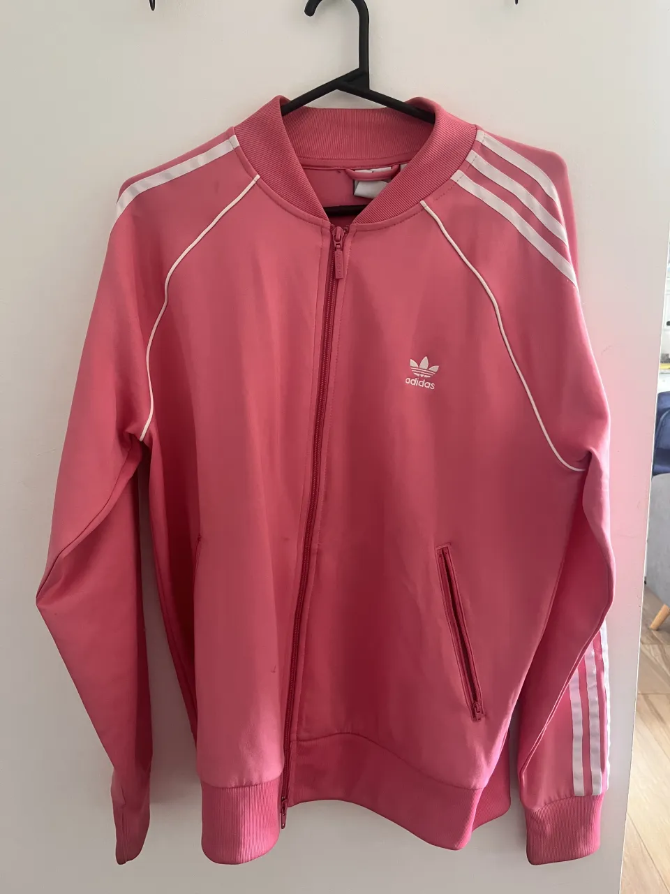 Campera deportiva rosa con cierre completo y cuello redondo. Posee tres tiras blancas en las mangas y detalles blancos en el cuello y puños. Tiene bolsillos laterales con cierre.