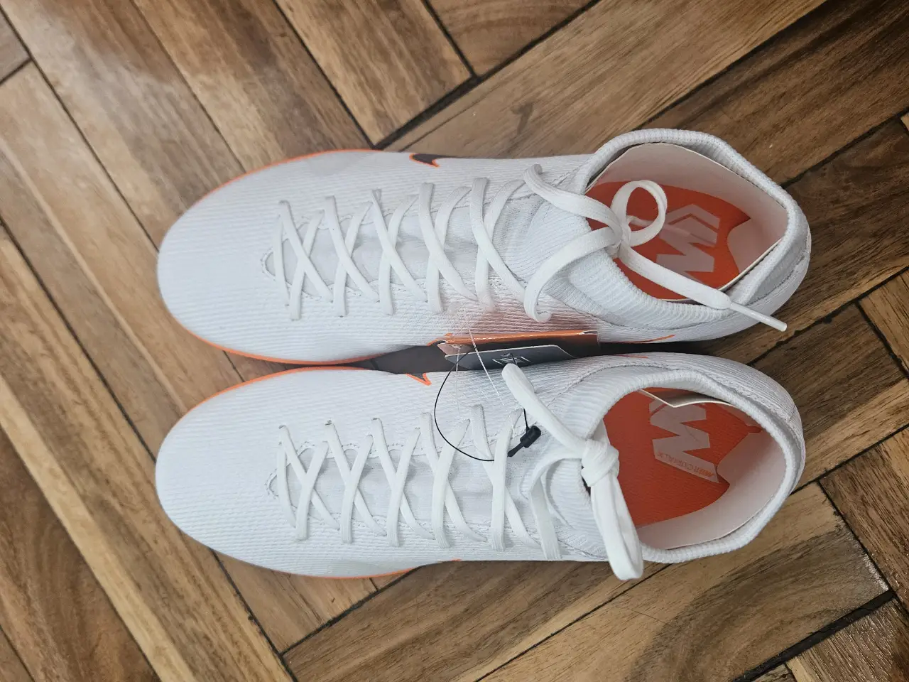 Zapatillas de fútbol Nike Mercurial, blancas con detalles en naranja y el logo en gris. Ideales para jugar en cancha indoor.