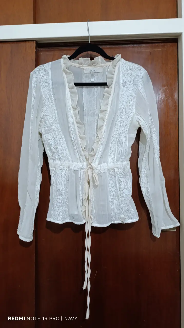Blusa de encaje blanca con detalles de volantes en el cuello y cierre con lazo. Ideal para un look romántico y elegante.