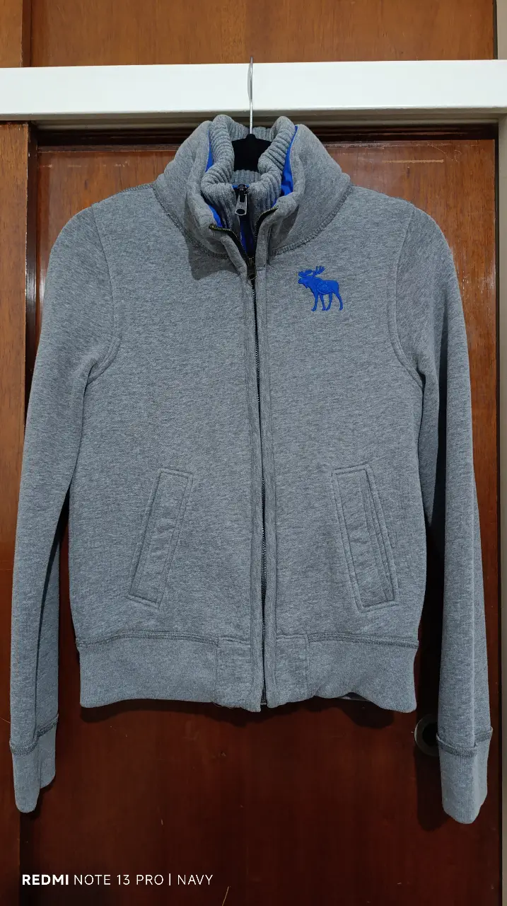 Buzo con cierre Abercrombie & Fitch para adolescente, color gris con detalles en azul. Con logo bordado en el pecho y forro interior acolchado. Ideal para un look casual y abrigado. Talle de adolescente.