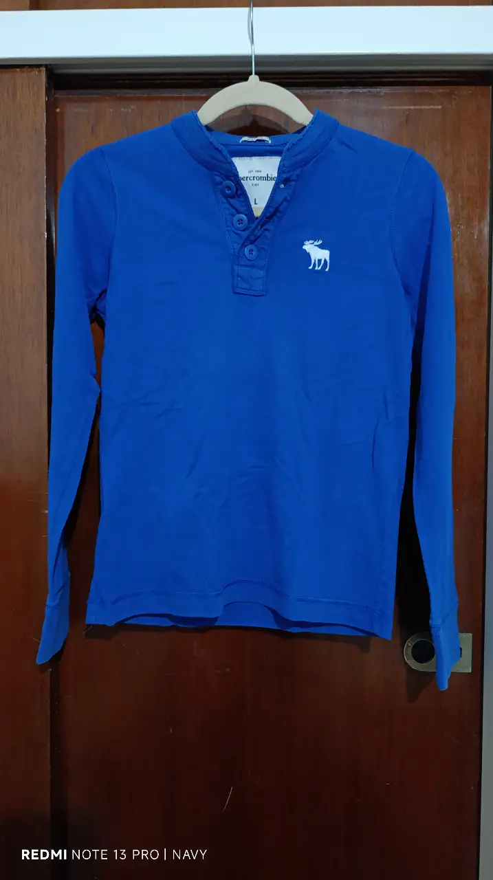 Remera manga larga azul de la marca Abercrombie Kids, con cuello mao y botones. Tiene el logo de un alce bordado en blanco en el pecho y la marca estampada en blanco en la manga.