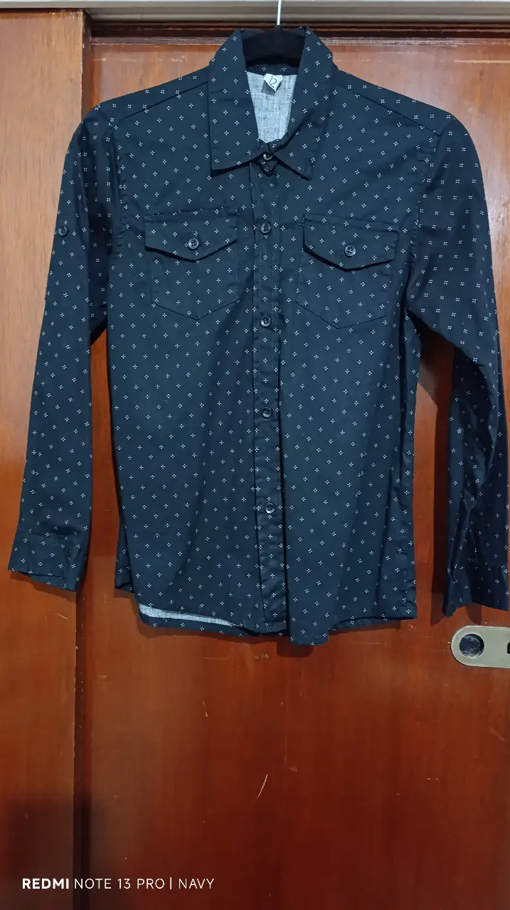 Camisa de niño negra con estampado de cruces blancas. Cuello camisero, dos bolsillos delanteros y cierre con botones. Ideal para un look casual y moderno.