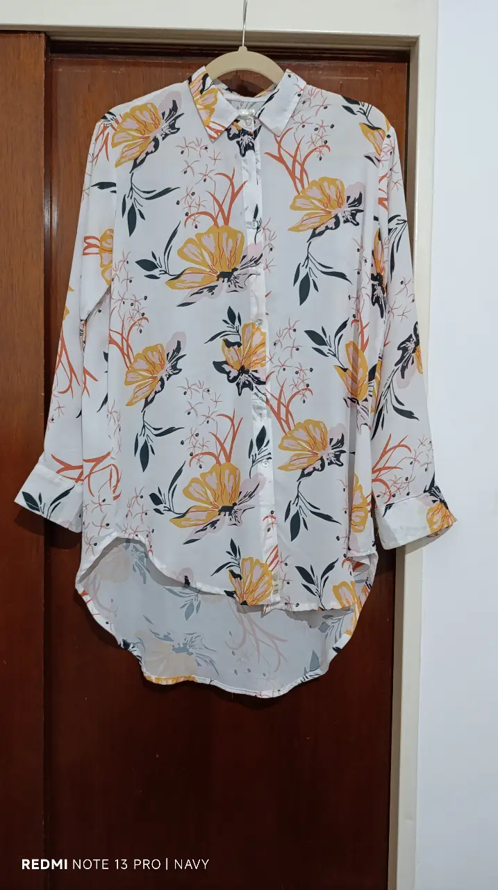 Blusa de mujer con estampado floral en tonos amarillos y detalles en negro y naranja sobre fondo blanco. Manga larga con puños y cuello camisero. Ideal para un look casual y fresco.