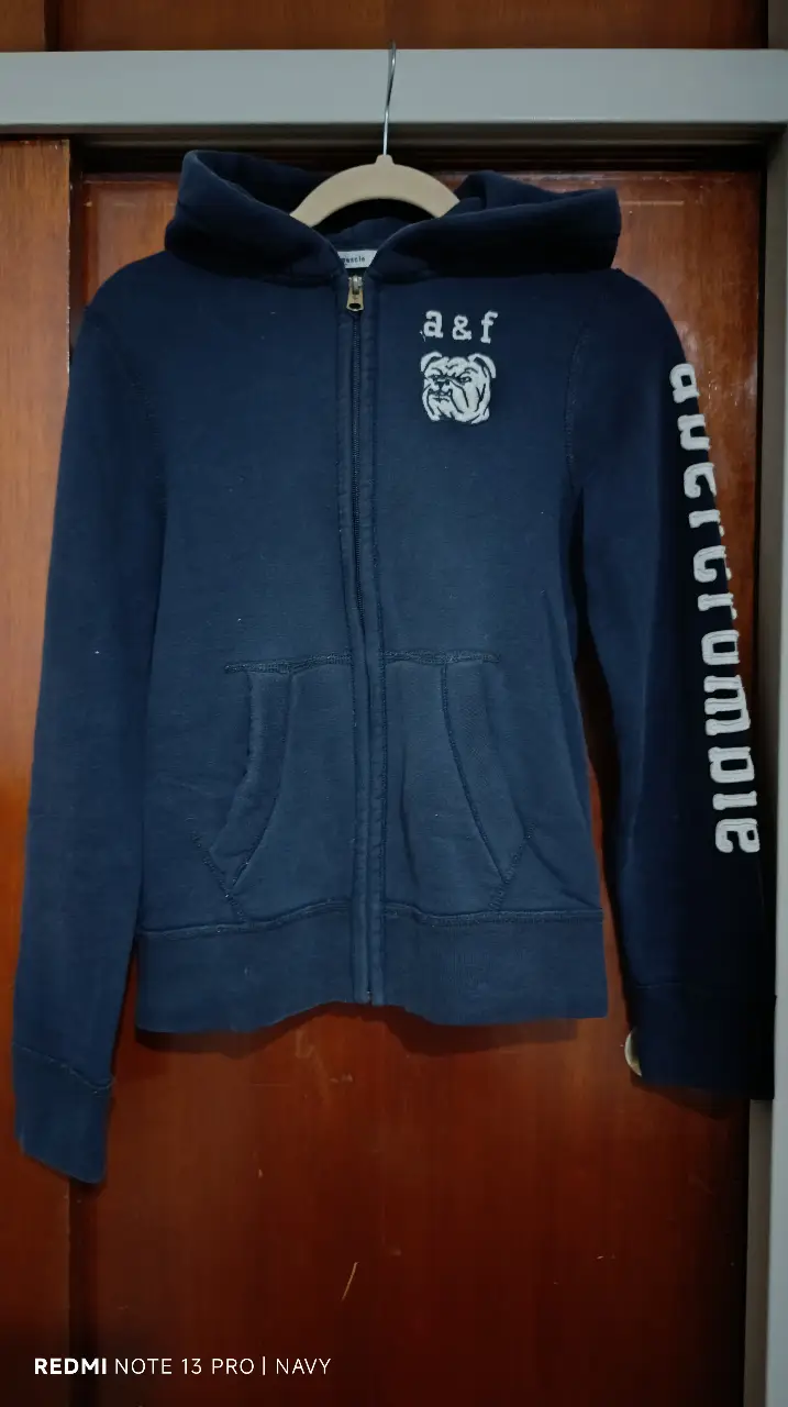 Buzo con cierre y capucha Abercrombie & Fitch para niño/adolescente, talle L. Color negro con logo de la marca en el pecho y detalle en la manga. Ideal para un look casual y cómodo.