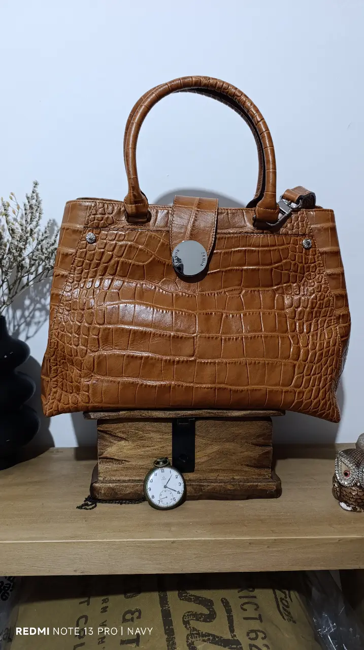 Cartera de mano PRÜNE de cuero marrón con textura de cocodrilo. Diseño elegante con manijas superiores y correa ajustable. Interior espacioso con compartimentos y cierre. Incluye bolsa protectora.