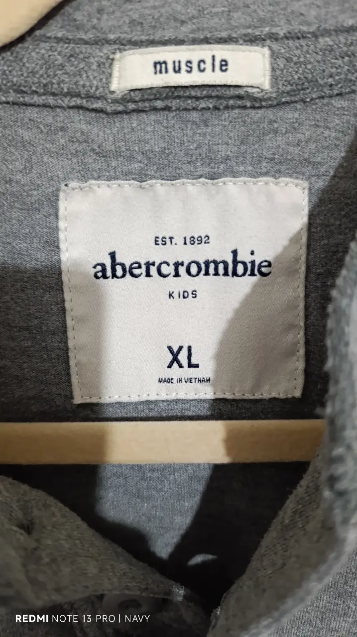 Remera manga larga gris de niño/adolescente con cuello alto y botones. Detalles en azul marino en las mangas y logo "A&F ATHL DEPT". Talle XL.