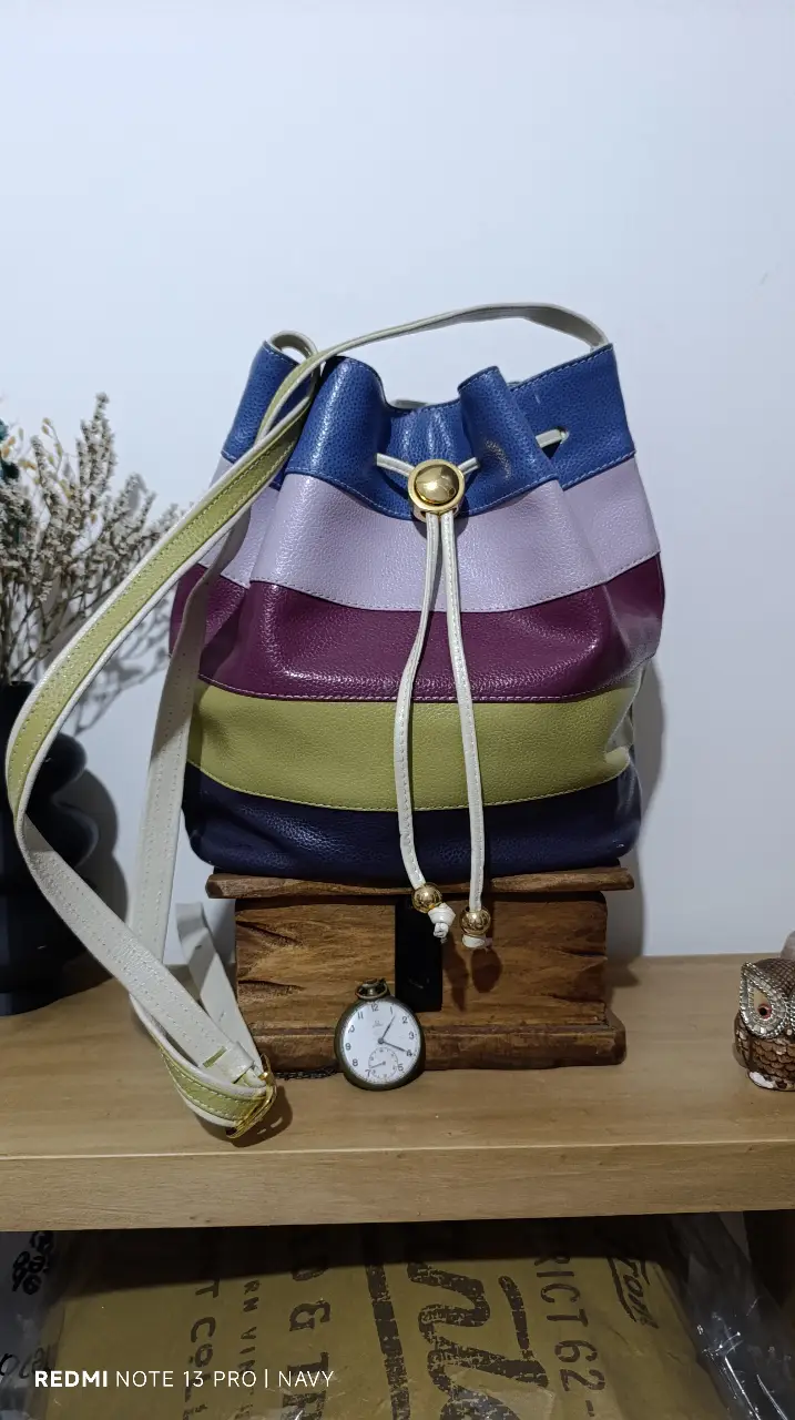 Cartera tipo balde de cuero, con diseño a rayas en varios colores. Cuenta con correa para colgar al hombro y cierre con cordón. Ideal para un look casual y moderno.