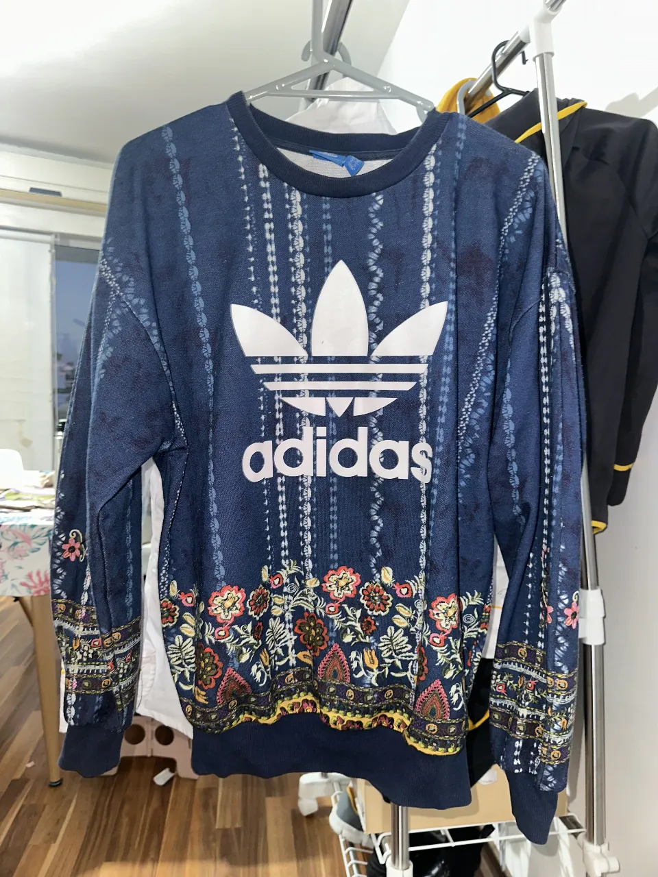 Buzo de cuello redondo Adidas con estampado floral en la parte inferior y en las mangas. El logo de Adidas en blanco en el pecho. Ideal para un look casual y cómodo.