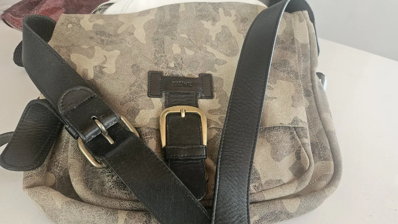 Bolso de hombro con estampa camuflada en tonos tierra y negro. Posee correa regulable con hebilla dorada y detalles en cuero negro.