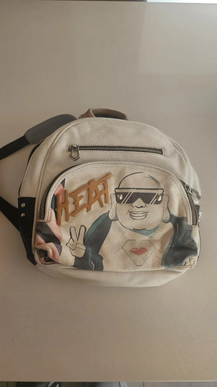 Mochila blanca con estampa de un personaje con anteojos y la palabra "HEAT". Tiene un bolsillo frontal con cierre y otro bolsillo superior con cierre. Las correas son de cuero blanco y gris claro. El interior es de tela blanca con estampa de corazones y tréboles.